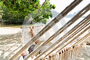 Young blonde woman relaxing in white hammock at Krabi Railey beach overlooking the harbour and mountains, Thailand