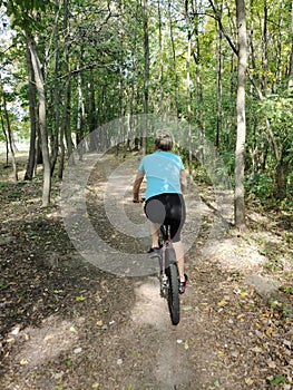 A young blonde woman rides a bicycle in the park on a path. Rear view. Tourism, sports and outdoor recreation