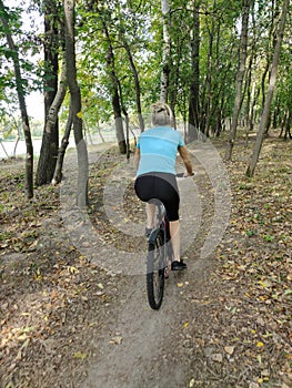 A young blonde woman rides a bicycle in the park on a path. Rear view. Tourism, sports and outdoor recreation