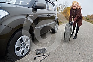 A young blonde woman rolls spare tire near her car with a flat tire