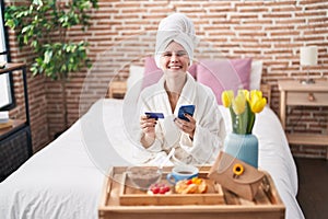 Young blonde woman shopping with smartphone and holding credit card having breakfast at bedroom