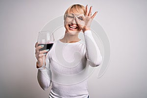 Young blonde woman with short hair drinking a glass of red wine over isolated background with happy face smiling doing ok sign