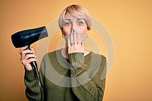 Young blonde woman with short hair drying her hair using hairdryer over yellow background cover mouth with hand shocked with shame