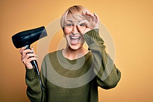 Young blonde woman with short hair drying her hair using hairdryer over yellow background with happy face smiling doing ok sign