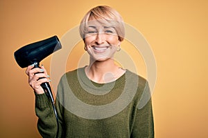 Young blonde woman with short hair drying her hair using hairdryer over yellow background with a happy face standing and smiling