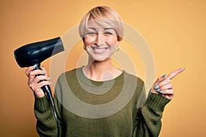 Young blonde woman with short hair drying her hair using hairdryer over yellow background very happy pointing with hand and finger