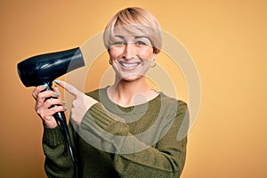 Young blonde woman with short hair drying her hair using hairdryer over yellow background very happy pointing with hand and finger