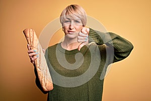 Young blonde woman with short hair holding fresh bread baguette over yellow background with angry face, negative sign showing