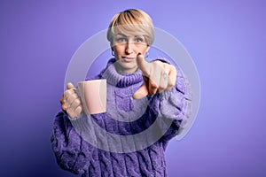 Young blonde woman with short hair wearing winter sweater drinking a cup of hot coffee pointing with finger to the camera and to