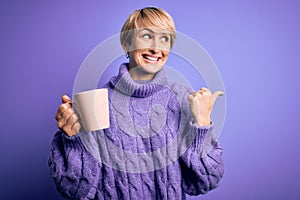 Young blonde woman with short hair wearing winter sweater drinking a cup of hot coffee pointing and showing with thumb up to the