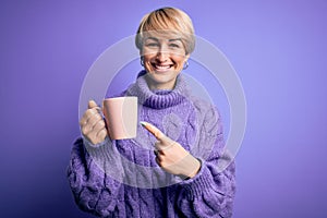 Young blonde woman with short hair wearing winter sweater drinking a cup of hot coffee very happy pointing with hand and finger