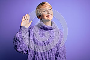 Young blonde woman with short hair wearing winter turtleneck sweater over purple background Waiving saying hello happy and