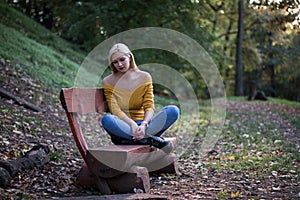Young blonde woman sitting alone on a wooden bench in the forest, sad and lonely