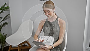 Young blonde woman sitting on chair using touchpad and earphones at waiting room