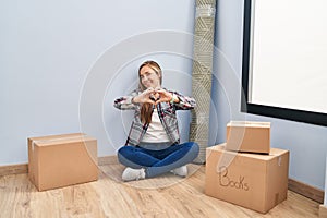 Young blonde woman sitting on the floor moving to a new home smiling in love doing heart symbol shape with hands