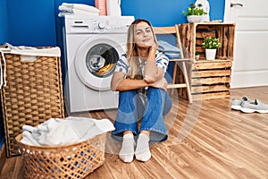 Young blonde woman sitting on the floor waiting for washing machine at laundry room