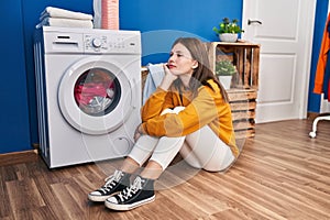 Young blonde woman sitting on floor waiting for washing machine at laundry room