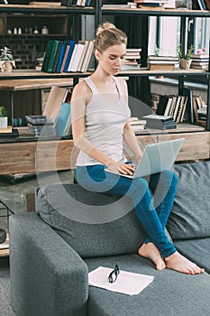 Young blonde woman sitting on sofa and using laptop