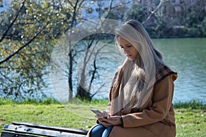 Young blonde woman, sitting on top of a bench, in a park, consulting social networks on her cell phone. Concept smartphone, social