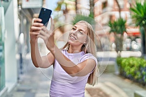 Young blonde woman smiling confident making selfie by the smartphone at street