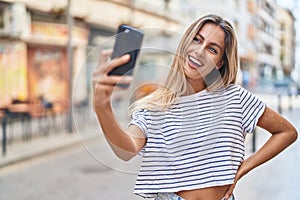 Young blonde woman smiling confident making selfie by the smartphone at street