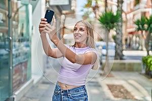 Young blonde woman smiling confident making selfie by the smartphone at street