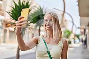 Young blonde woman smiling confident making selfie by the smartphone at street