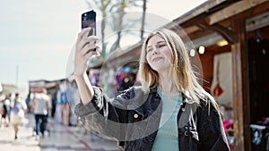 Young blonde woman smiling confident making selfie by the smartphone at street market