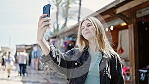 Young blonde woman smiling confident making selfie by the smartphone at street market