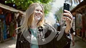 Young blonde woman smiling confident making selfie by the smartphone at street market