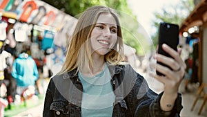 Young blonde woman smiling confident making selfie by the smartphone at street market