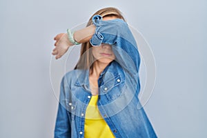 Young blonde woman standing over blue background covering eyes with arm, looking serious and sad