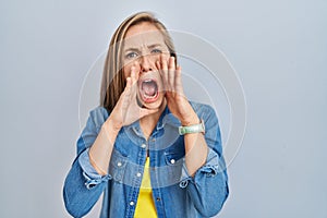 Young blonde woman standing over blue background shouting angry out loud with hands over mouth