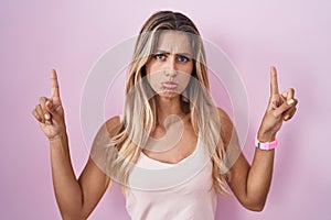 Young blonde woman standing over pink background pointing up looking sad and upset, indicating direction with fingers, unhappy and