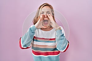 Young blonde woman standing over pink background shouting angry out loud with hands over mouth