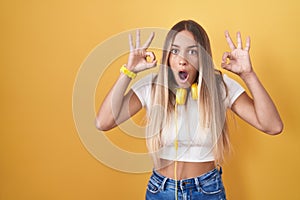 Young blonde woman standing over yellow background wearing headphones looking surprised and shocked doing ok approval symbol with