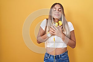 Young blonde woman standing over yellow background wearing headphones smiling with hands on chest with closed eyes and grateful