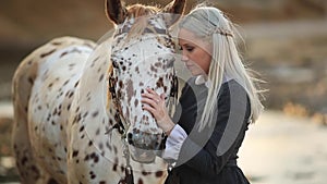 Young Blonde woman stroking and hugging horse. Beautiful lady with her white stallion on seashore enjoying nature. Love and