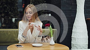 Young blonde woman takes a cup of coffee to her lips sitting at the table in cafe