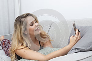 young blonde woman taking photos with her smart phone on the sofa in her living room