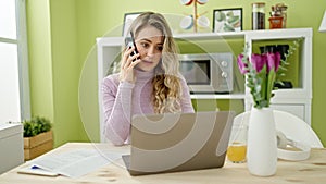 Young blonde woman talking on smartphone using laptop at dinning room