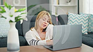Young blonde woman talking on smartphone using laptop at home