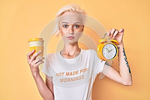 Young blonde woman with tattoo holding alarm clock and take away cup of coffee relaxed with serious expression on face