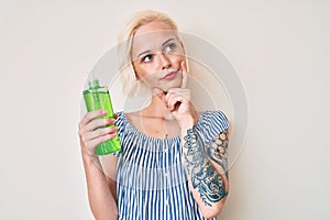 Young blonde woman with tattoo holding aloe vera serious face thinking about question with hand on chin, thoughtful about