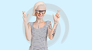 Young blonde woman with tattoo wearing casual clothes and glasses gesturing finger crossed smiling with hope and eyes closed