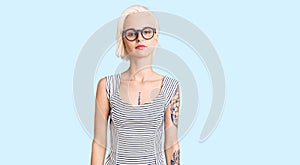 Young blonde woman with tattoo wearing casual clothes and glasses relaxed with serious expression on face