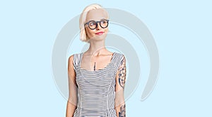 Young blonde woman with tattoo wearing casual clothes and glasses smiling looking to the side and staring away thinking