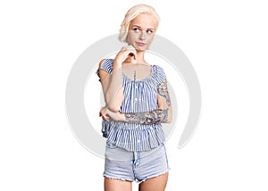 Young blonde woman with tattoo wearing casual clothes serious face thinking about question with hand on chin, thoughtful about