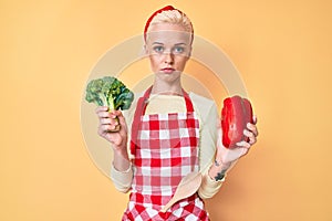 Young blonde woman with tattoo wearing cook apron holding broccoli and red pepper relaxed with serious expression on face