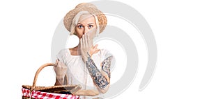 Young blonde woman with tattoo wearing summer hat and holding picnic wicker basket with bread covering mouth with hand, shocked